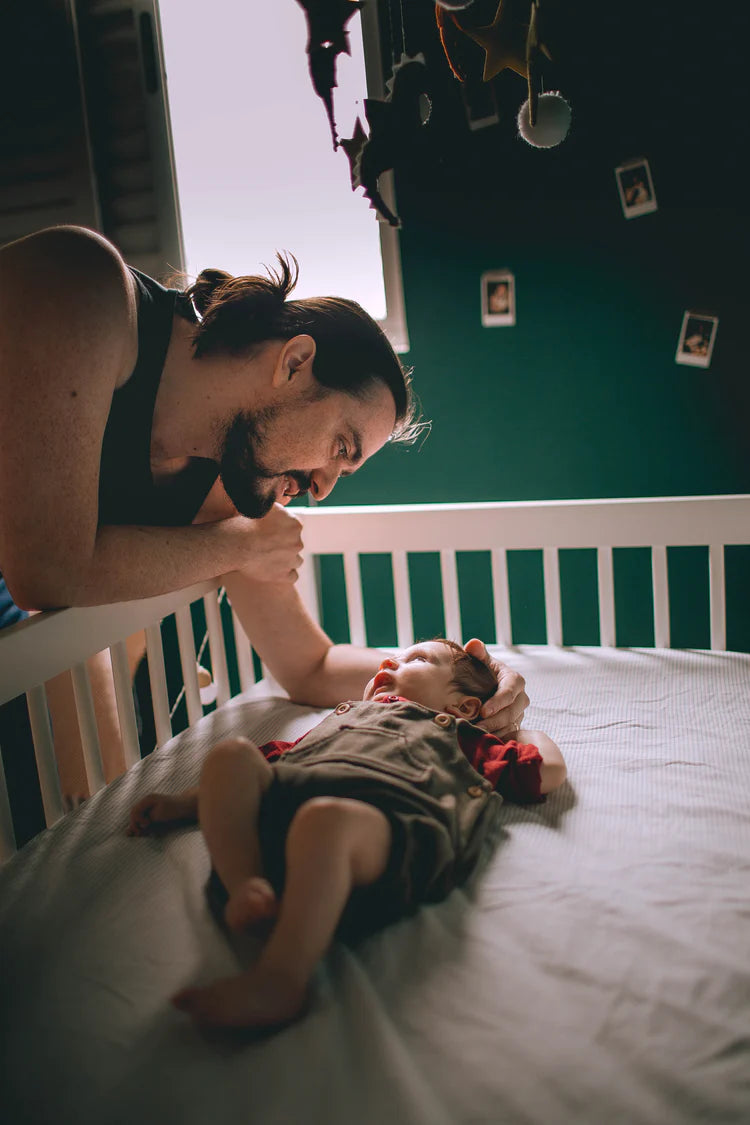 Junger Vater beugt sich über das Babybett – Thema ist rückenfreundliche Kinderpflege